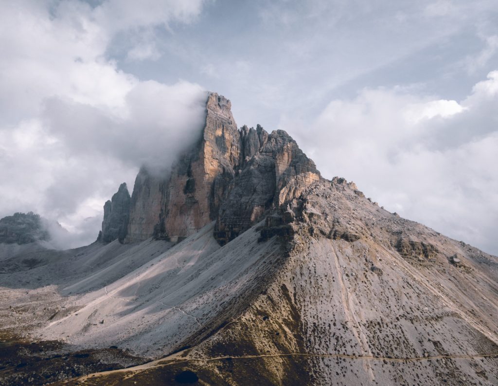Rocky mountain peaks rise sharply into a cloudy sky, with mist partially covering the tallest peak. The slopes below are rugged and mostly barren, with subtle trails winding through the landscape.