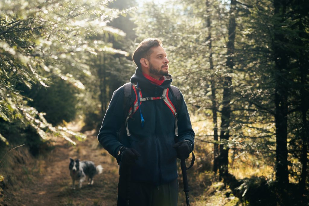 A man wearing outdoor hiking gear stands on a forest trail, looking to the side with sunlight filtering through the trees. A black and white dog walks behind him among the trees.
